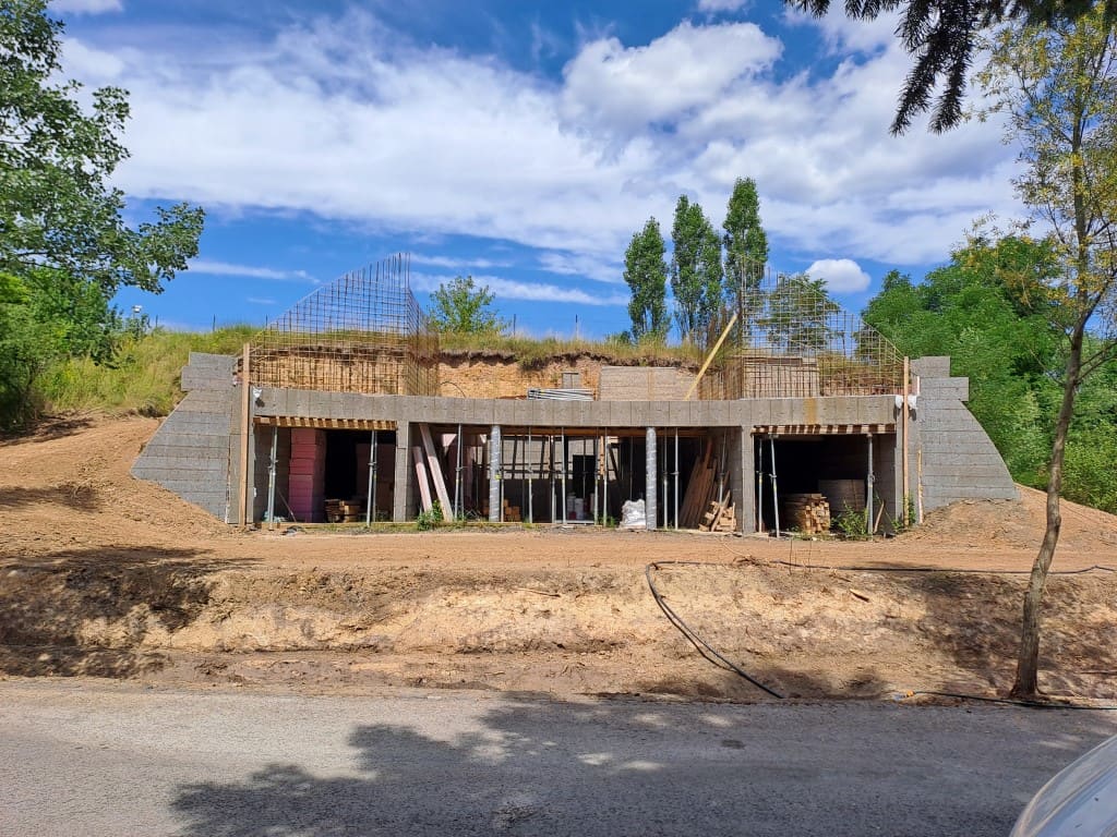 Earth-sheltered house under construction in a grassy hillside, concrete piers and rebar exposed