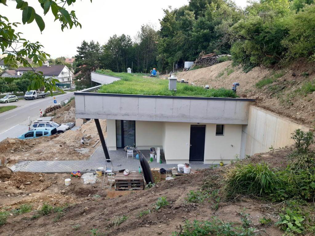 Earth-sheltered house with green roof and cream stucco facade nearing completion on a wooded hillside
