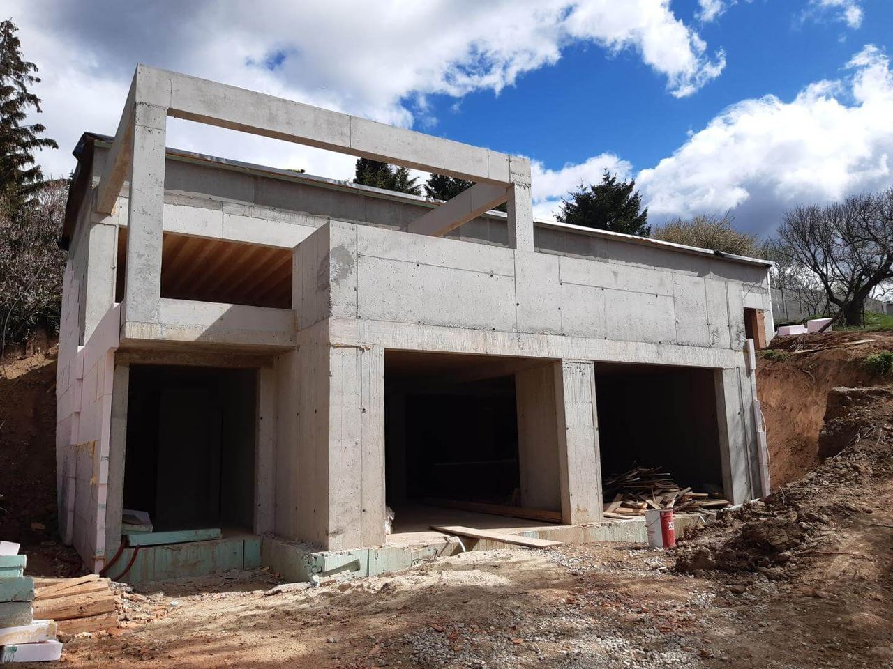 Raw concrete shell of an earth-sheltered house under construction, cantilevered upper floor, pink insulation, blue sky