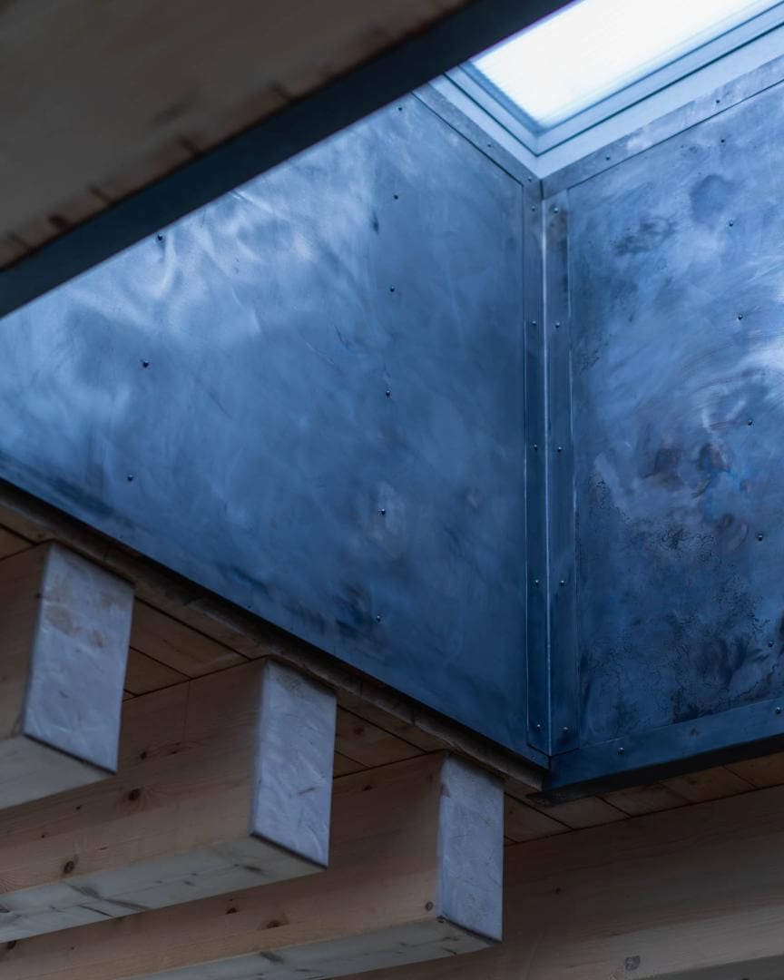 Riveted metal skylight shaft channels daylight through timber roof framing of an earth-sheltered house