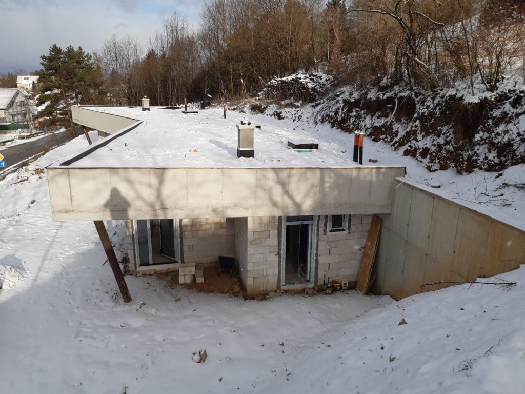 Snow-covered earth-sheltered house under construction, concrete roof and block walls on a tree-lined hillside