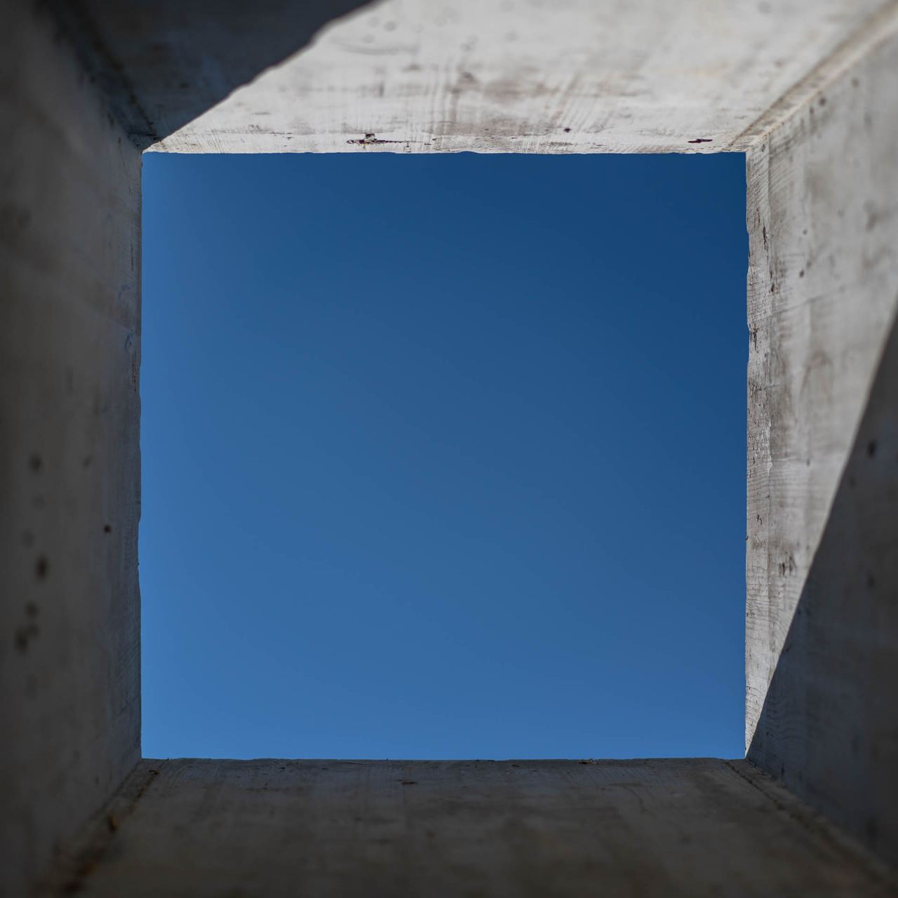 Board-formed concrete skylight opening framing blue sky with sharp diagonal sunlight shadow