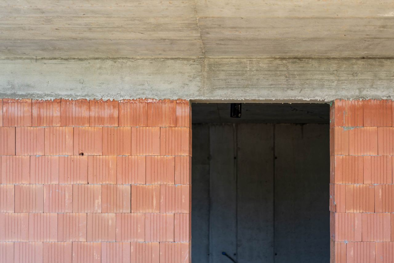 Close-up of hollow clay brick wall with concrete lintel band above a dark door opening, junction box visible