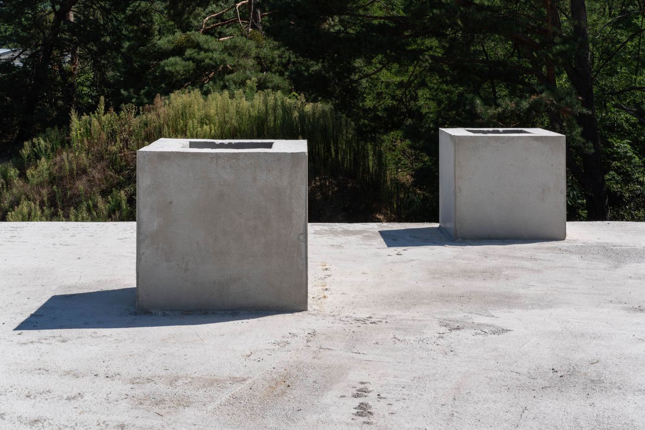 Two concrete cube shafts on a slab, forest backdrop, sunlit earth-sheltered house construction site
