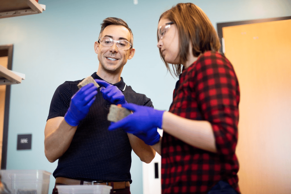 Two researchers in gloves and goggles examine sandy bio-based concrete samples in a lab